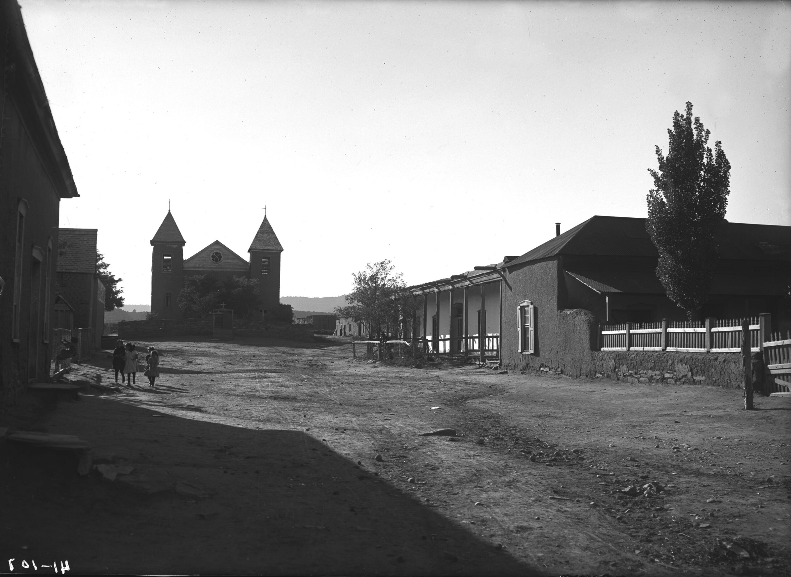 A black and white photo featuring a dirt road in the foreground, a church behind and businesses flanking.